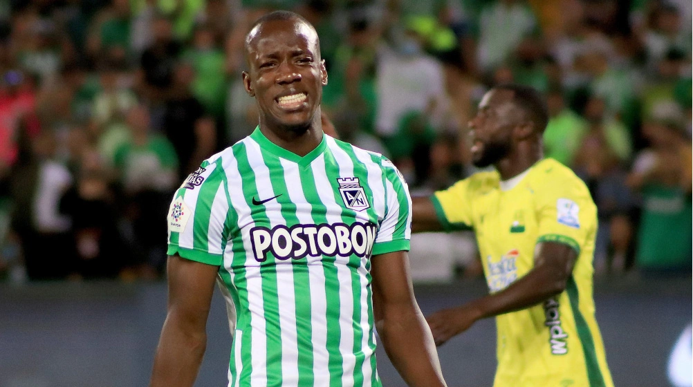 Jogador de futebol Baldomero Perlaza, vestindo o uniforme listrado verde e branco do Atlético Nacional, durante uma partida, com outro jogador desfocado ao fundo.